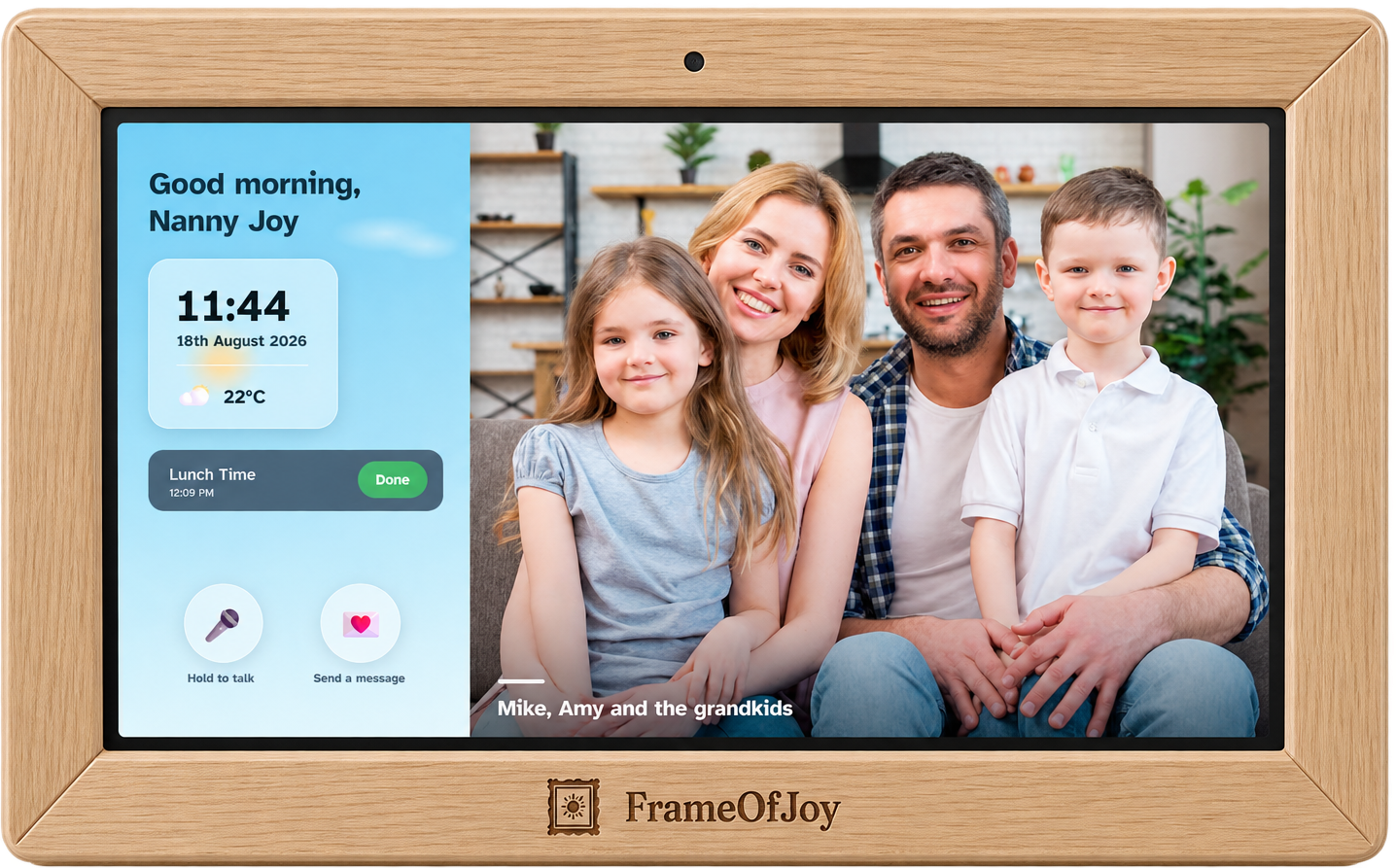 FrameOfJoy digital photo frame in a light wood surround, standing on its stand. The screen shows a good-morning greeting by name, the time, date and weather, a lunch reminder marked done, a family photo, and the hold-to-talk and send-a-message buttons.